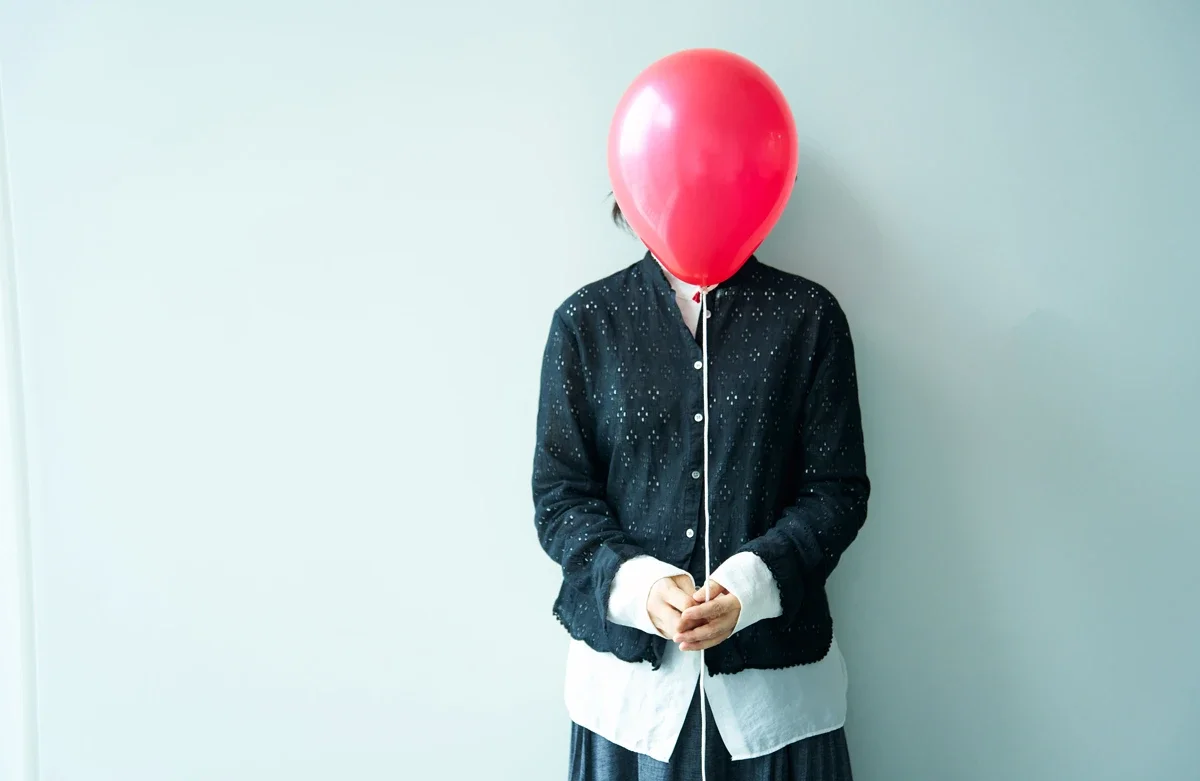 白い壁の前に立っている女性の顔に赤い風船が被さっている。