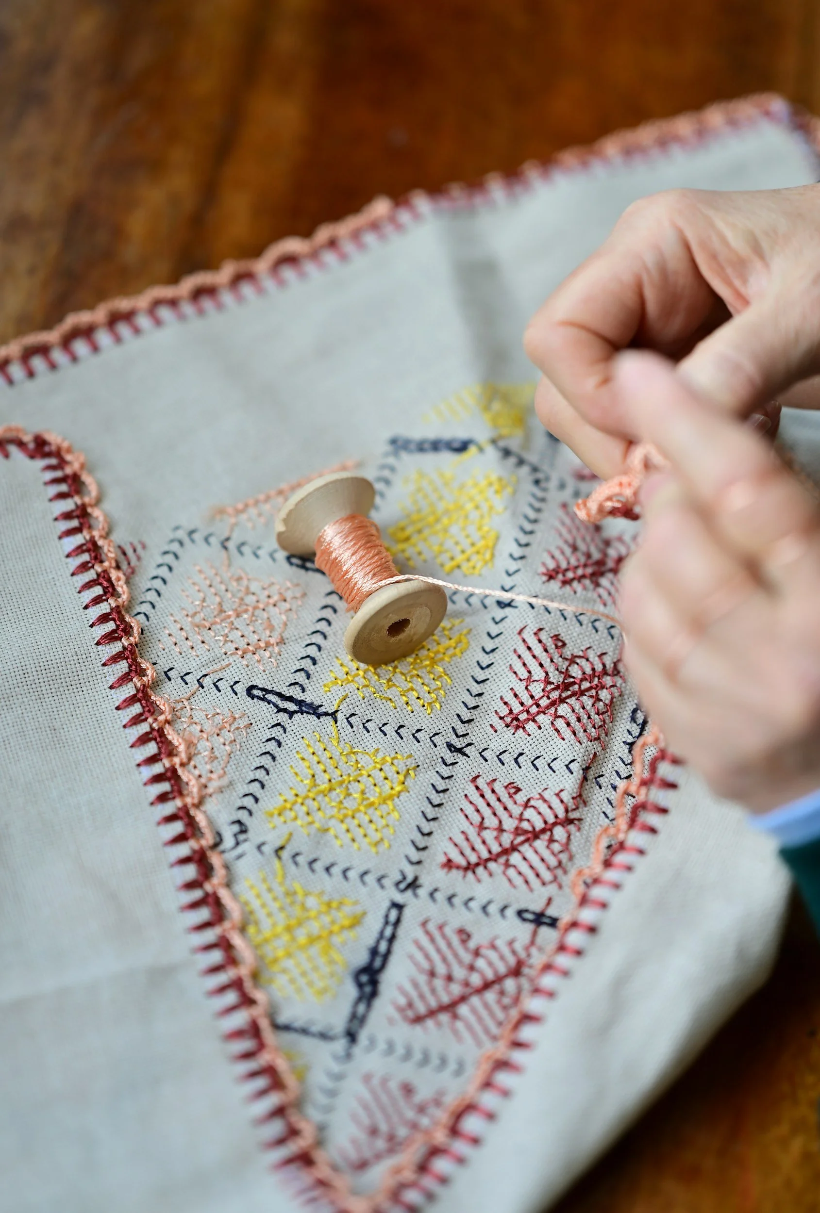 A close-up of a person's hand sewing colorful embroidery on fabric using a wooden spool of peach-colored thread. The fabric has a pattern of red, yellow, and blue stitched designs.
