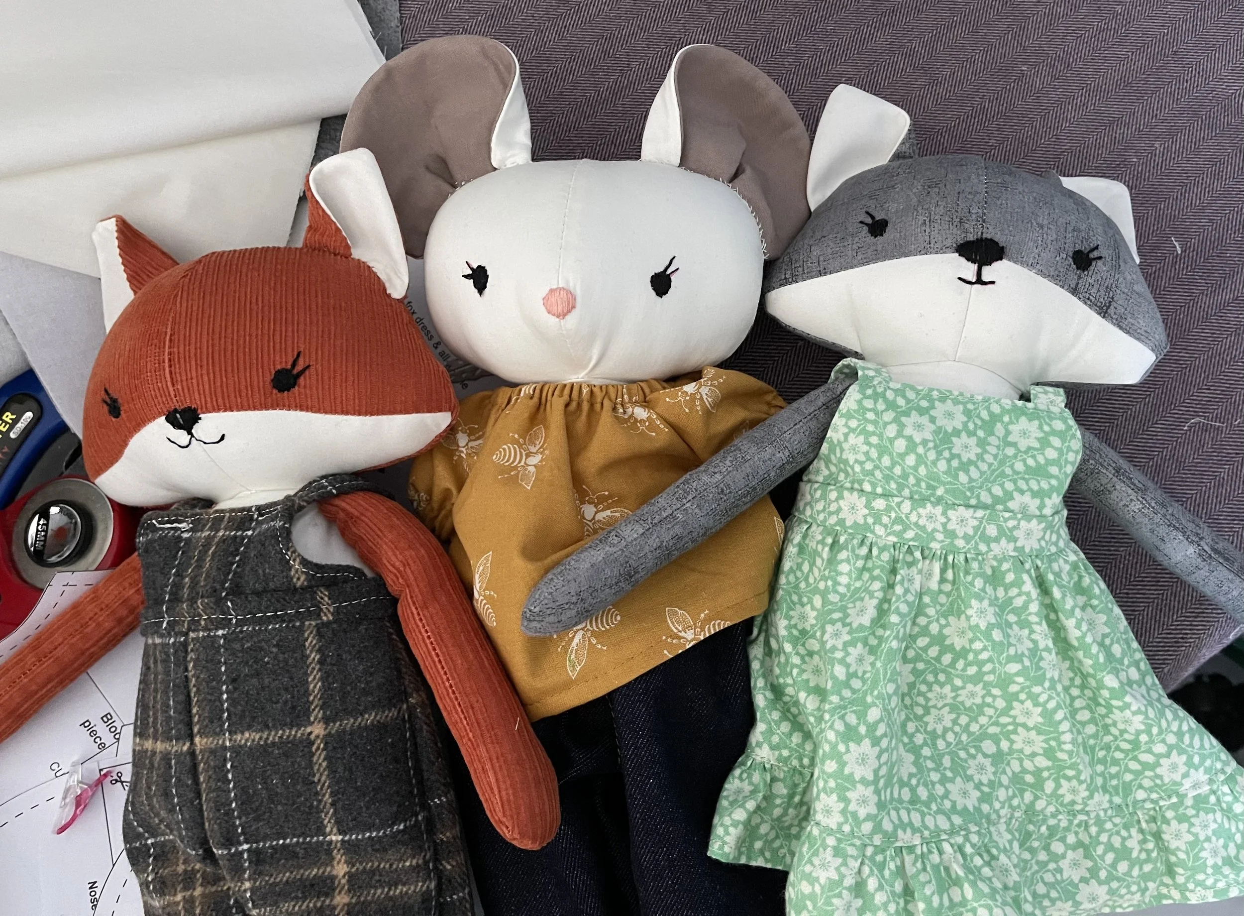 Three plush animal dolls with fabric bodies and embroidered faces. The first is an orange fox with plaid clothing, the second is a white bunny with gray ears and a mustard yellow dress, and the third is a gray wolf with white ears and a green floral dress. They are laying on a purple surface with other household items around.