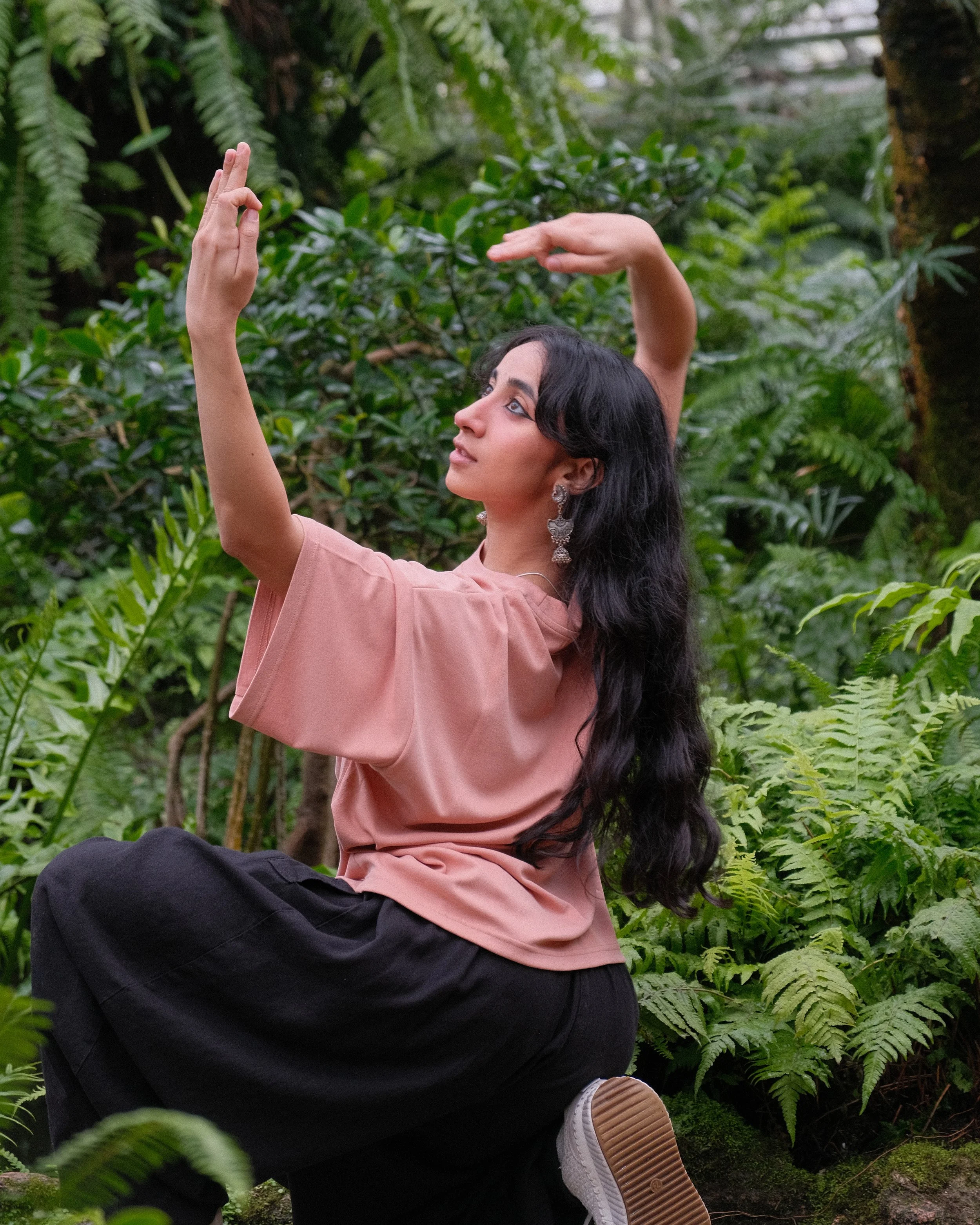 A woman with long black hair, wearing a pink top, black pants, and white sneakers, sits on the ground in a lush green forest, gesturing gracefully with her arms raised and looking to the side.