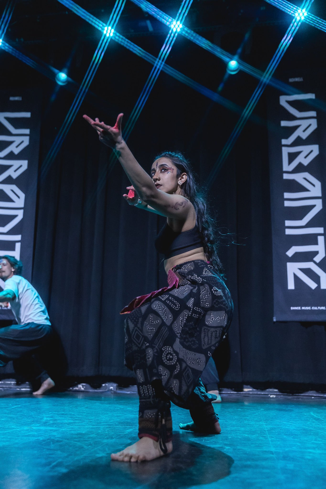 Performer in black crop top and patterned pants performing contemporary dance on stage with blue lighting and black curtains.