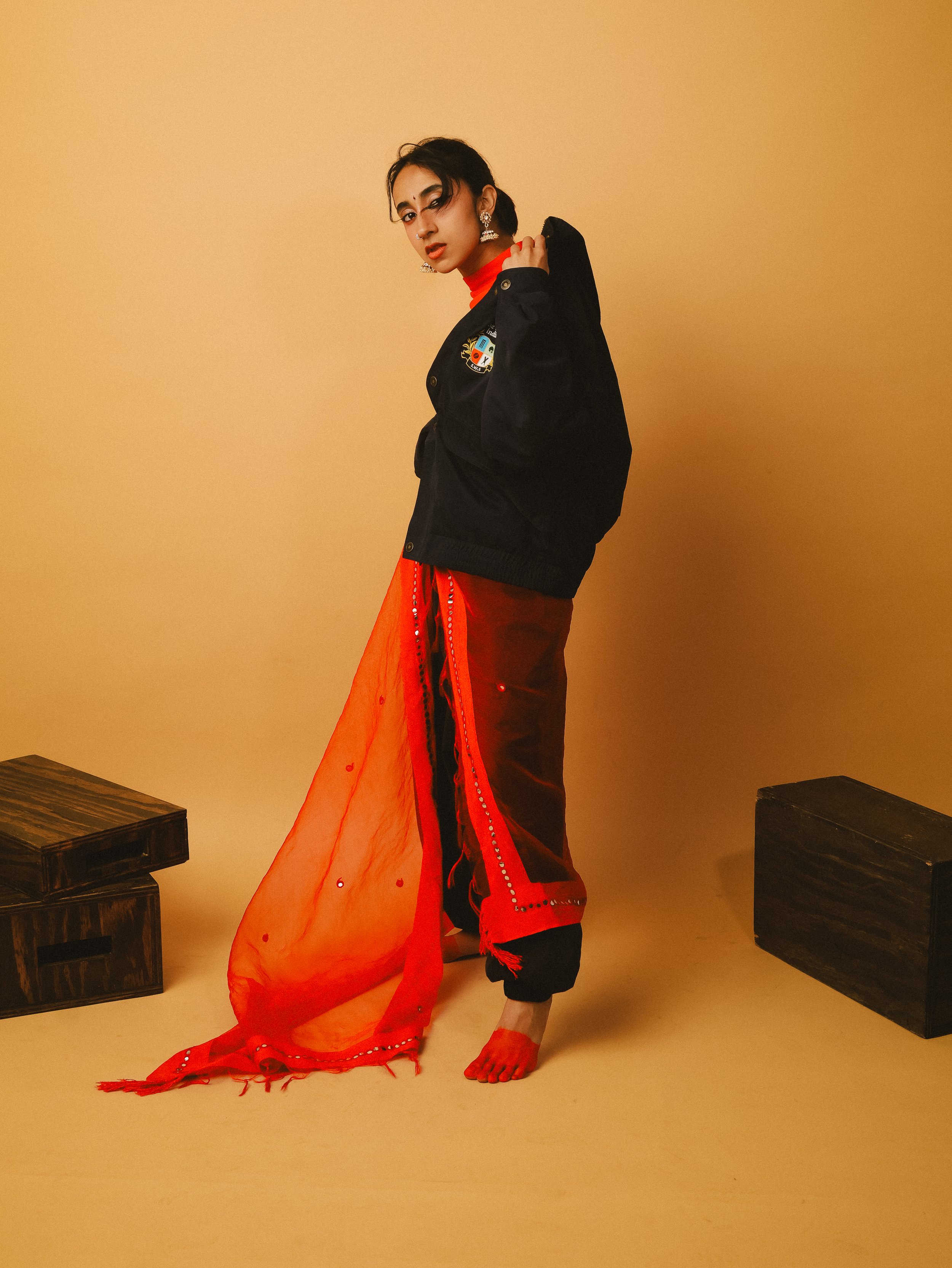 A woman standing barefoot in front of a beige background, dressed in a red and black outfit with a black jacket and sheer red fabric, posing with one hand on her shoulder and looking at the camera.