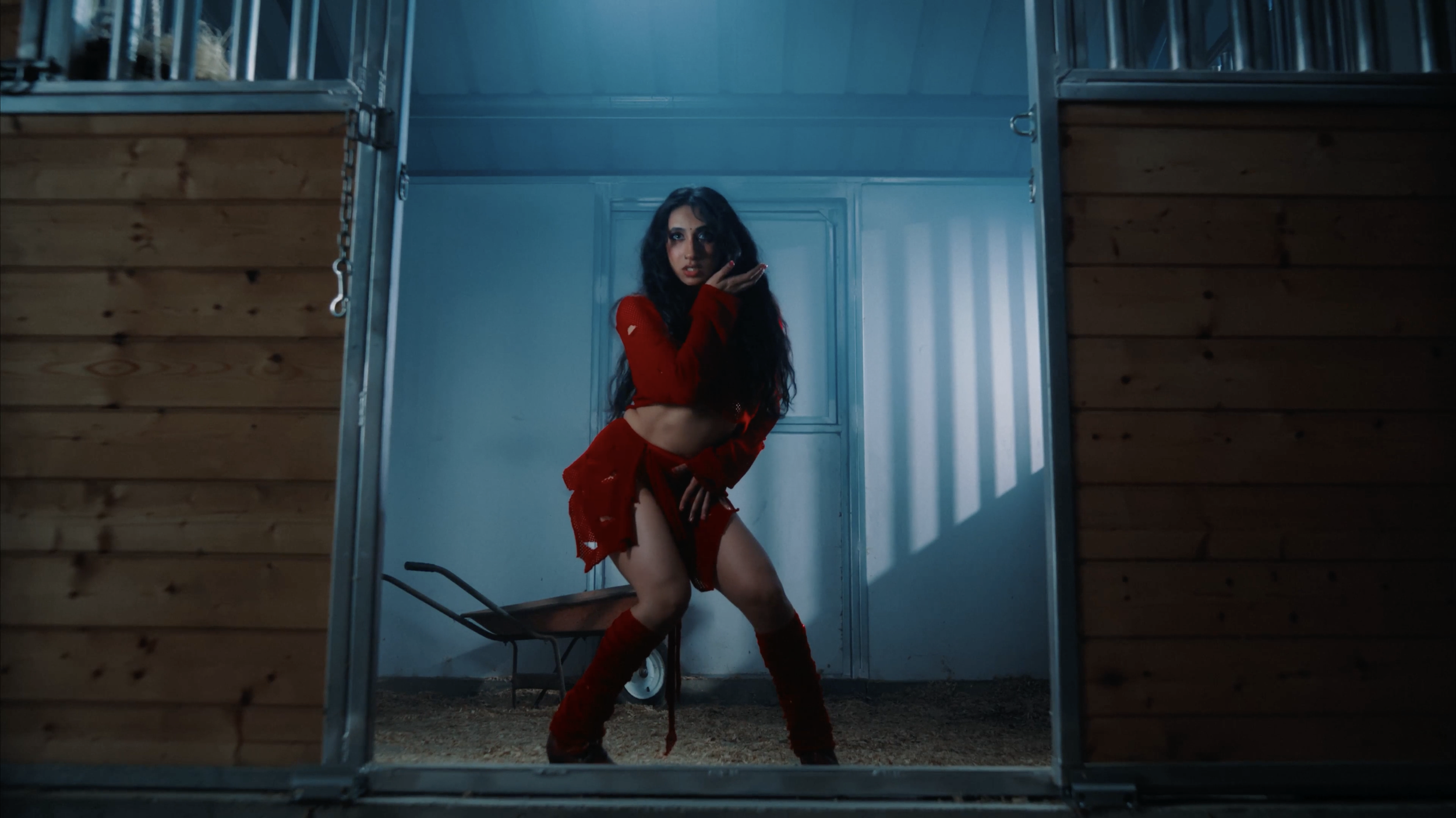 Woman in red outfit standing in a barn-like structure, seen through a window or opening.