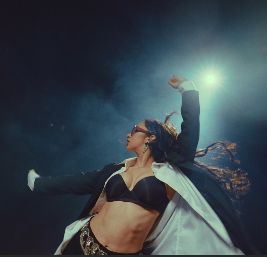 A woman performing on stage, wearing sunglasses, a black bra, a black and white blazer, and a decorative belt, with her arm raised and long hair flowing.