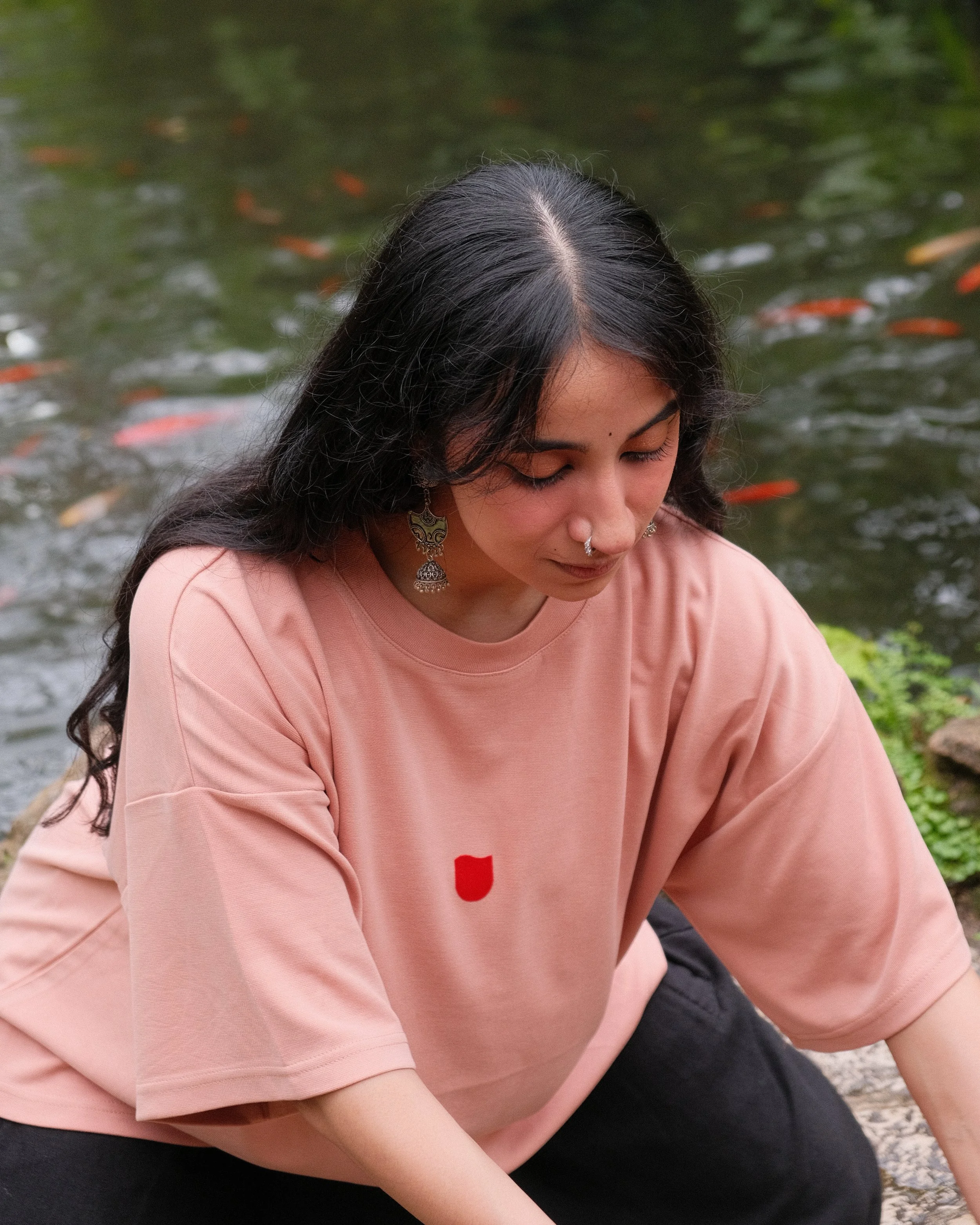 A woman with long black hair, wearing earrings, sitting near a pond with fish visible in the water.