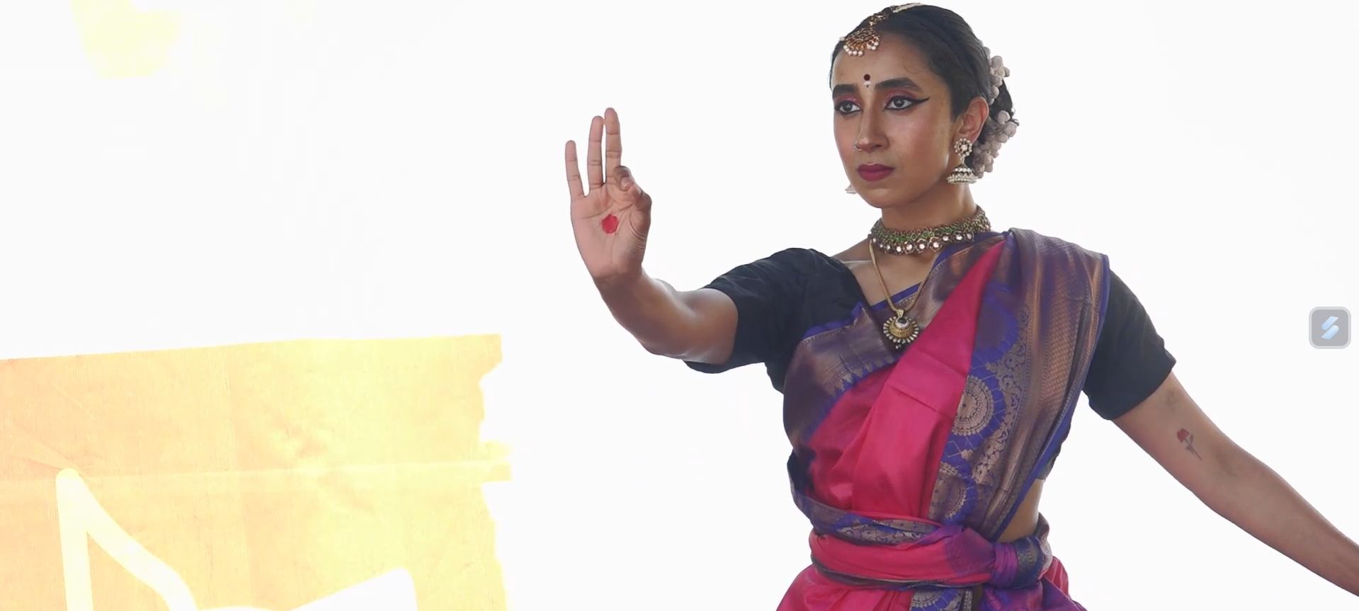 A young woman dressed in traditional Indian attire performing a classical dance. She wears a pink and purple saree, ornate jewelry including earrings, necklaces, and a headpiece. Her hair is decorated with flowers, and she has a small red bindi on her forehead. She is making a gesture with her right hand, against a plain light background.