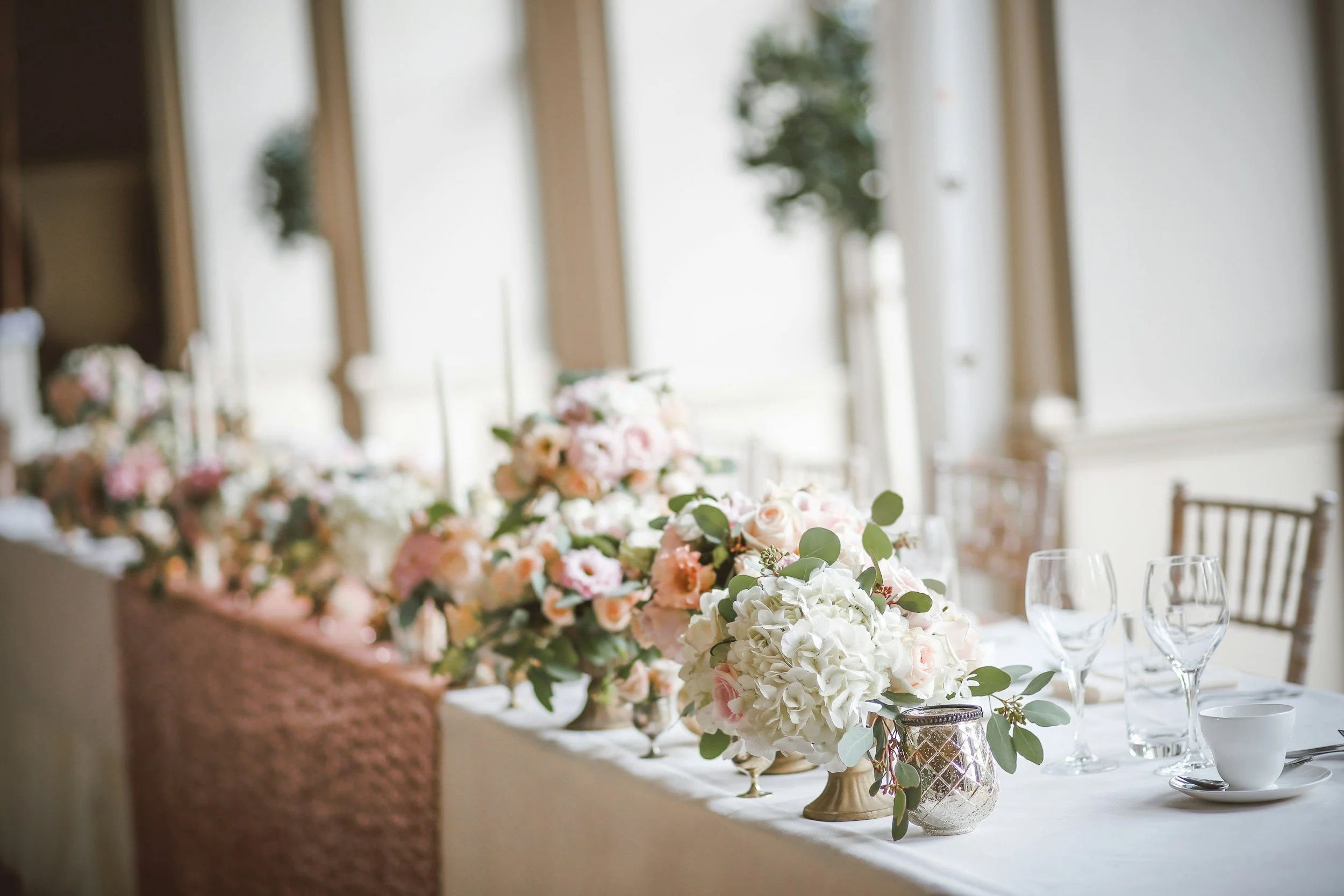 Elegant wedding reception table decorated with pink and white flowers, glassware, and candles in a bright room with large windows.