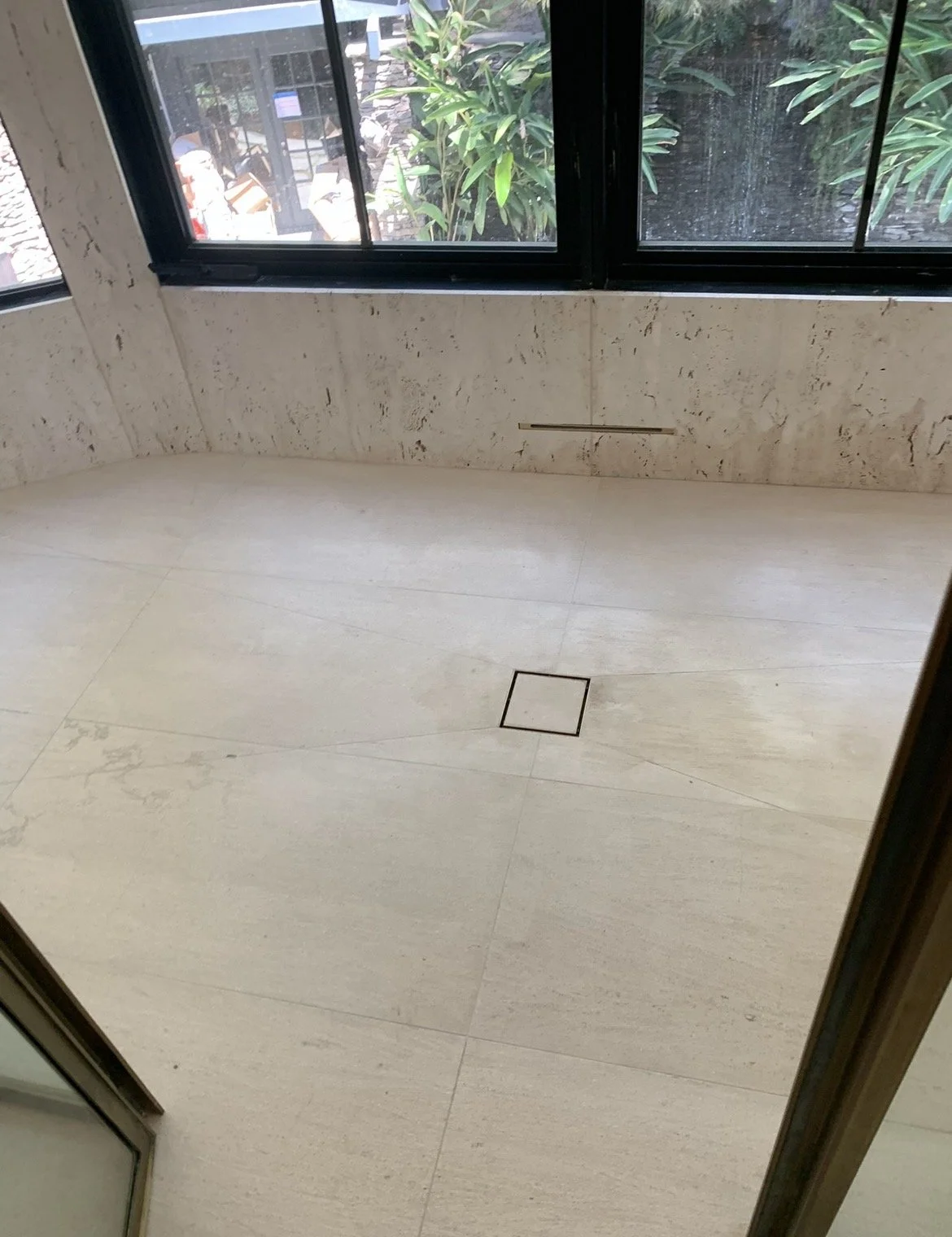 Empty room with cream-colored tiled floor, marble walls, black-framed windows showing greenery outside, and a floor drain.