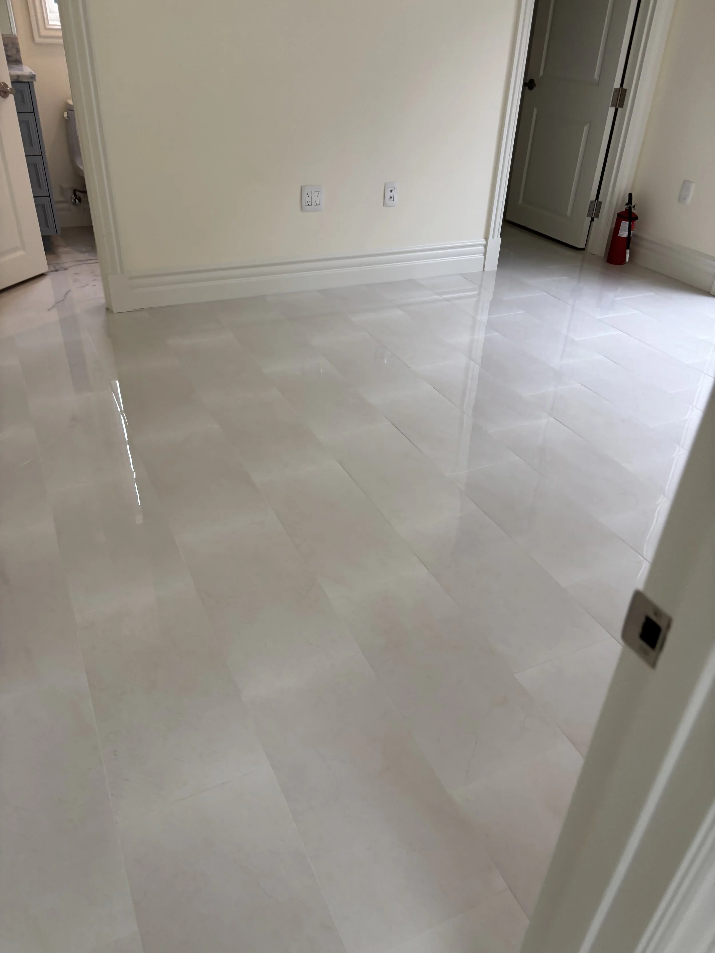 Empty room with shiny beige tile flooring, cream walls, a white door slightly open revealing a closet, a fire extinguisher near the door, and a glimpse into a bathroom with marble flooring.