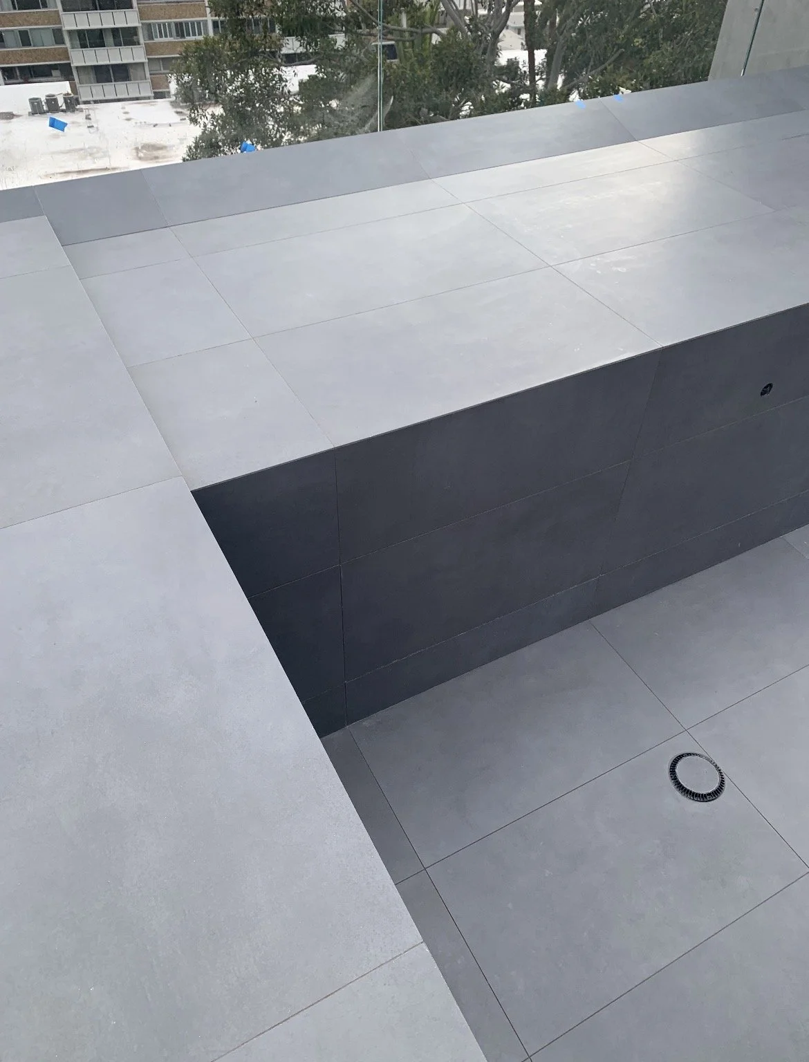 A modern indoor space with gray tiled flooring and a built-in rectangular black tub or bench, with a drain on the floor and a large glass window showing trees and buildings outside.