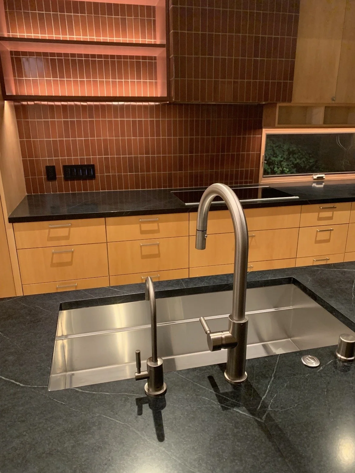 Modern kitchen with black countertop, stainless steel sink, and brass faucet. Brown tile backsplash, wooden cabinets, and a window showing greenery outside.