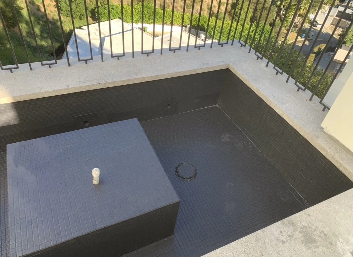Empty balcony with black-tiled hot tub or spa, with a drain and a small pipe, surrounded by a concrete railing with black metal bars, overlooking green trees and neighboring buildings.