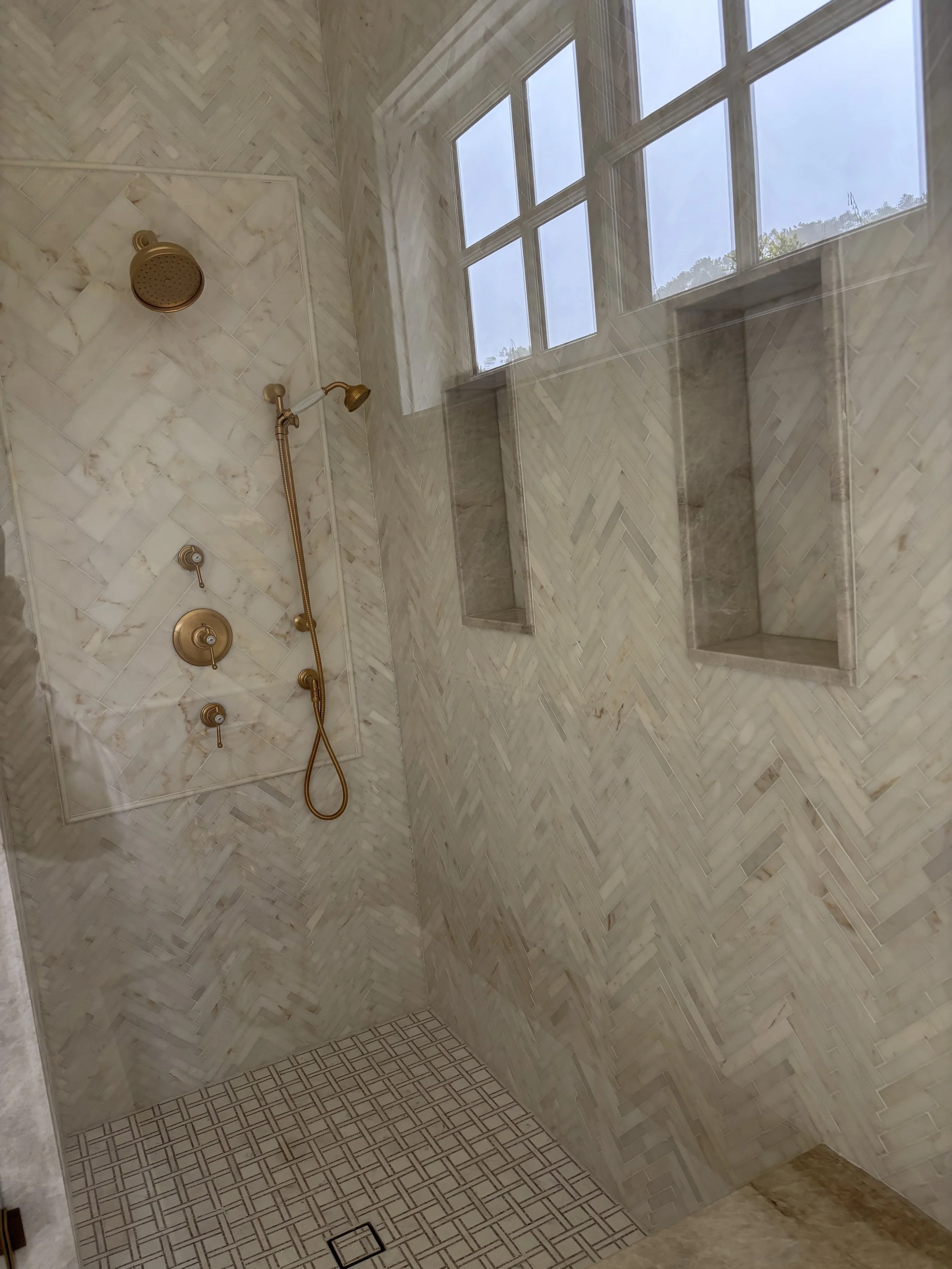 Shower area with marble walls, a window with multiple panes, and a gold-colored showerhead and fixtures.