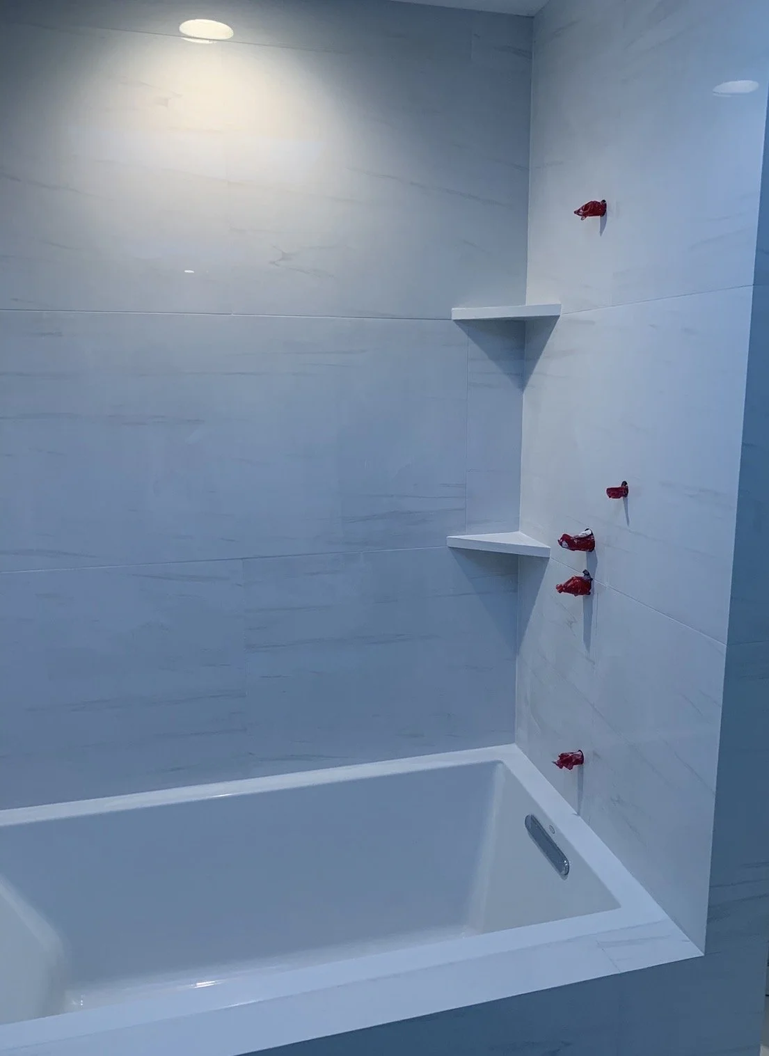 Empty bathtub in a bathroom with unfinished plumbing, with red caps on the water fixtures and white shelves on the tiled wall.