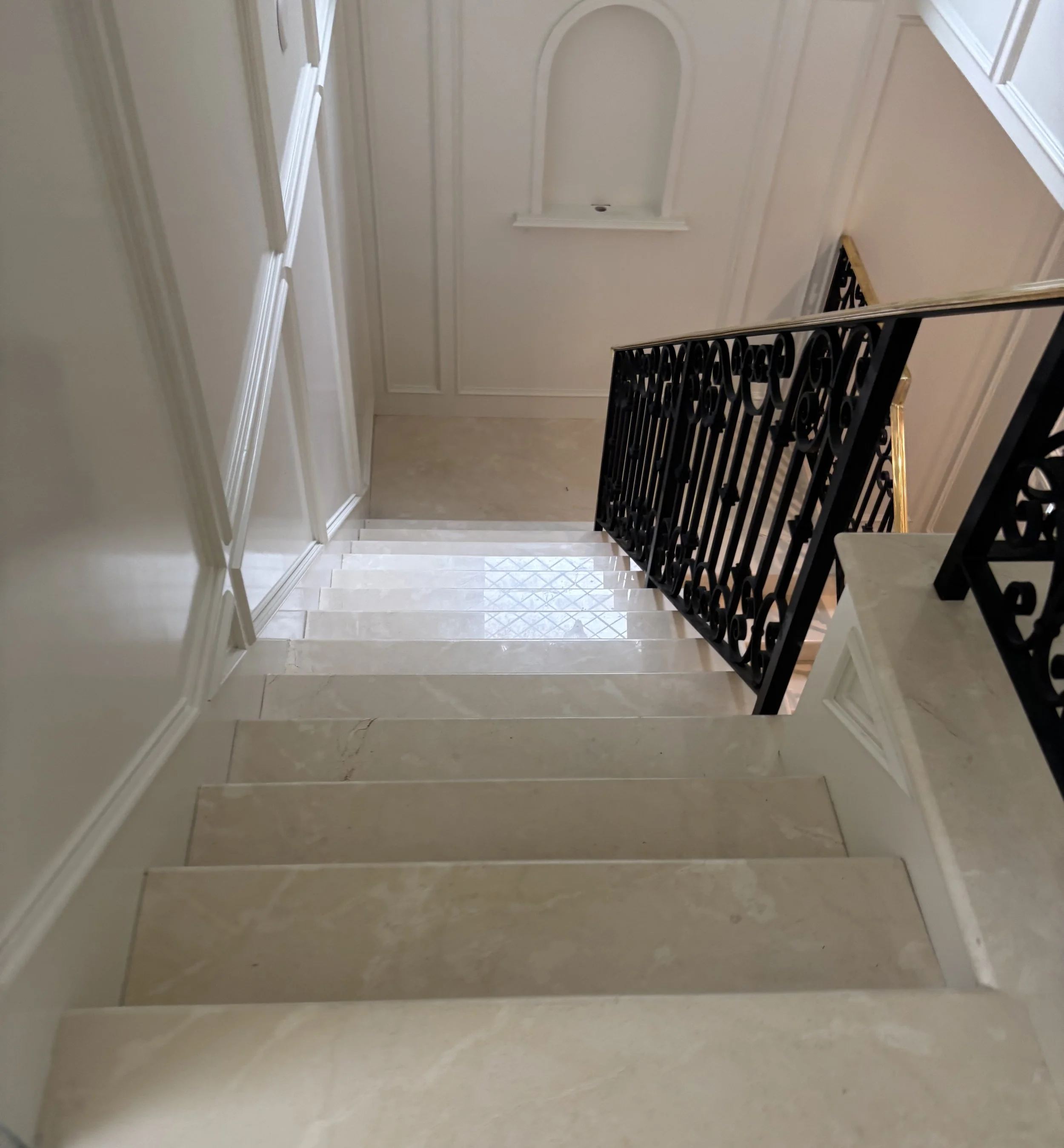 A staircase descending from an upper floor with beige marble steps and a decorative black metal railing, with a small wall niche in the background.