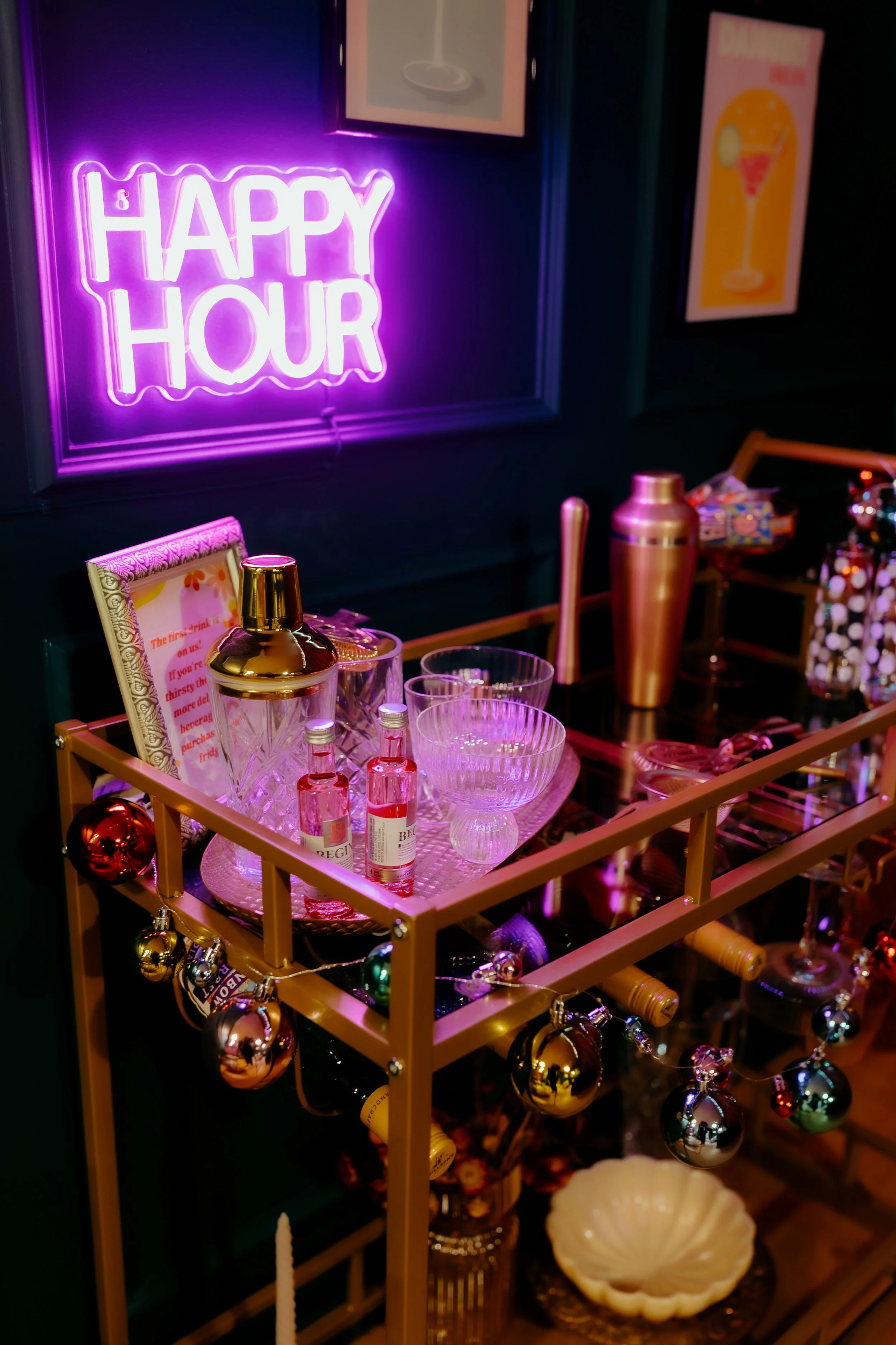 Bar cart decorated with Christmas ornaments, filled with glassware and bottles, illuminated by a pink "Happy Hour" neon sign, against a dark wall with colorful posters.