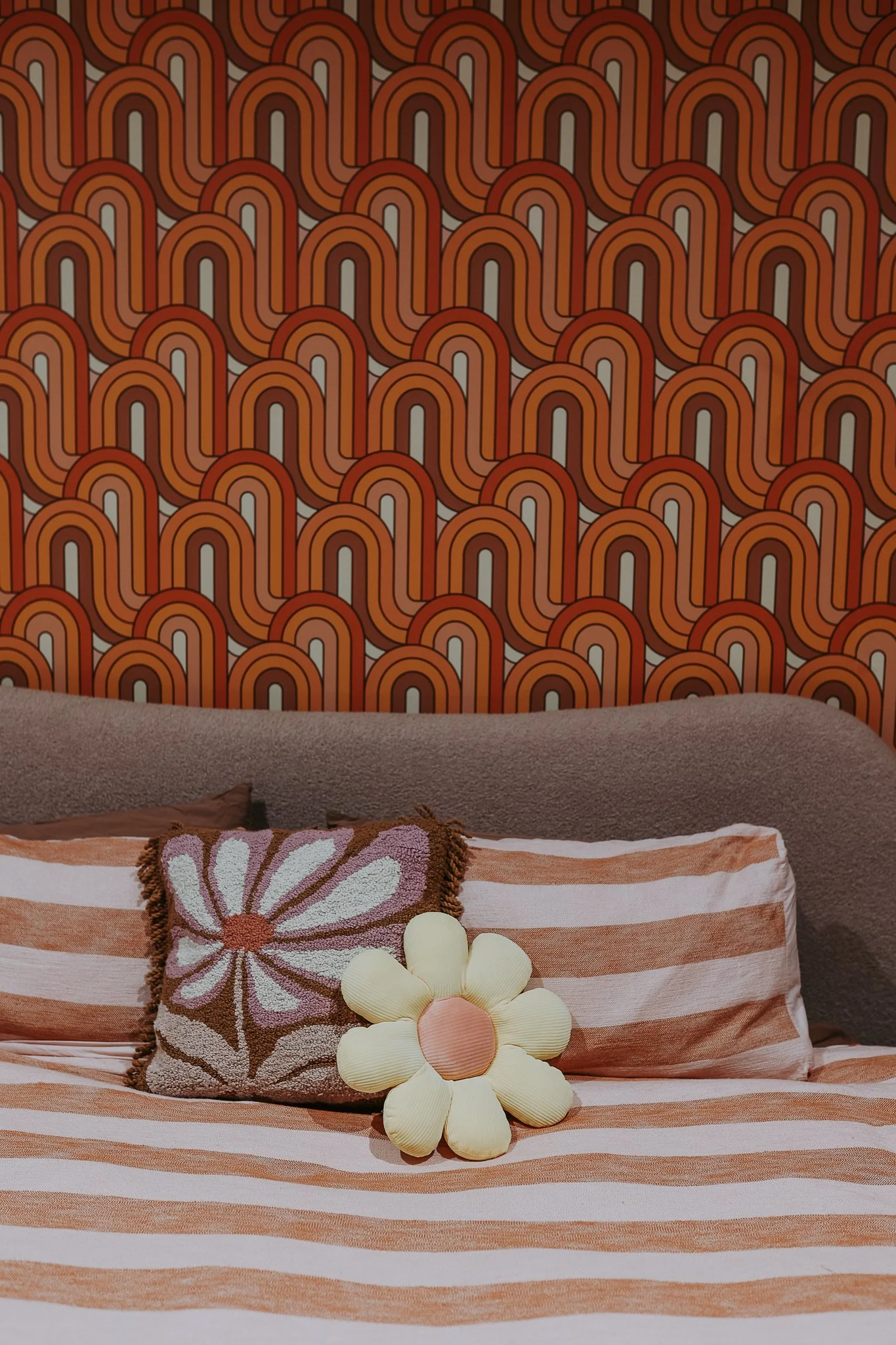 A bed with striped peach and white bedding, decorated with a pillow featuring a floral design and a plush flower accent, set against a retro patterned wallpaper with orange and brown rainbow shapes.