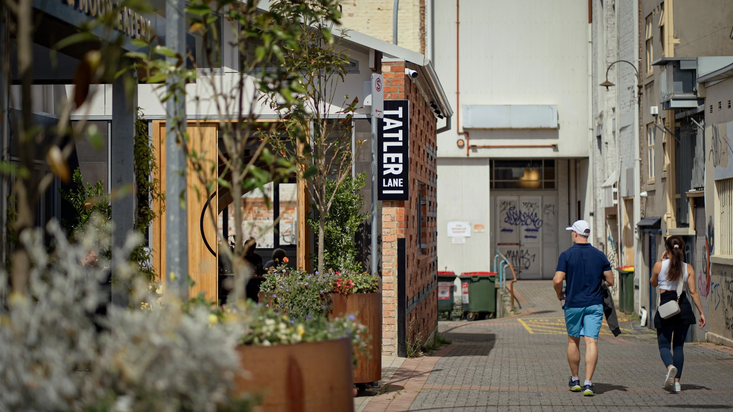 Two people, a man in a white cap and a woman, walk down a narrow alleyway with plants and graffiti on the walls. A sign reads 'TATLER LANE.'