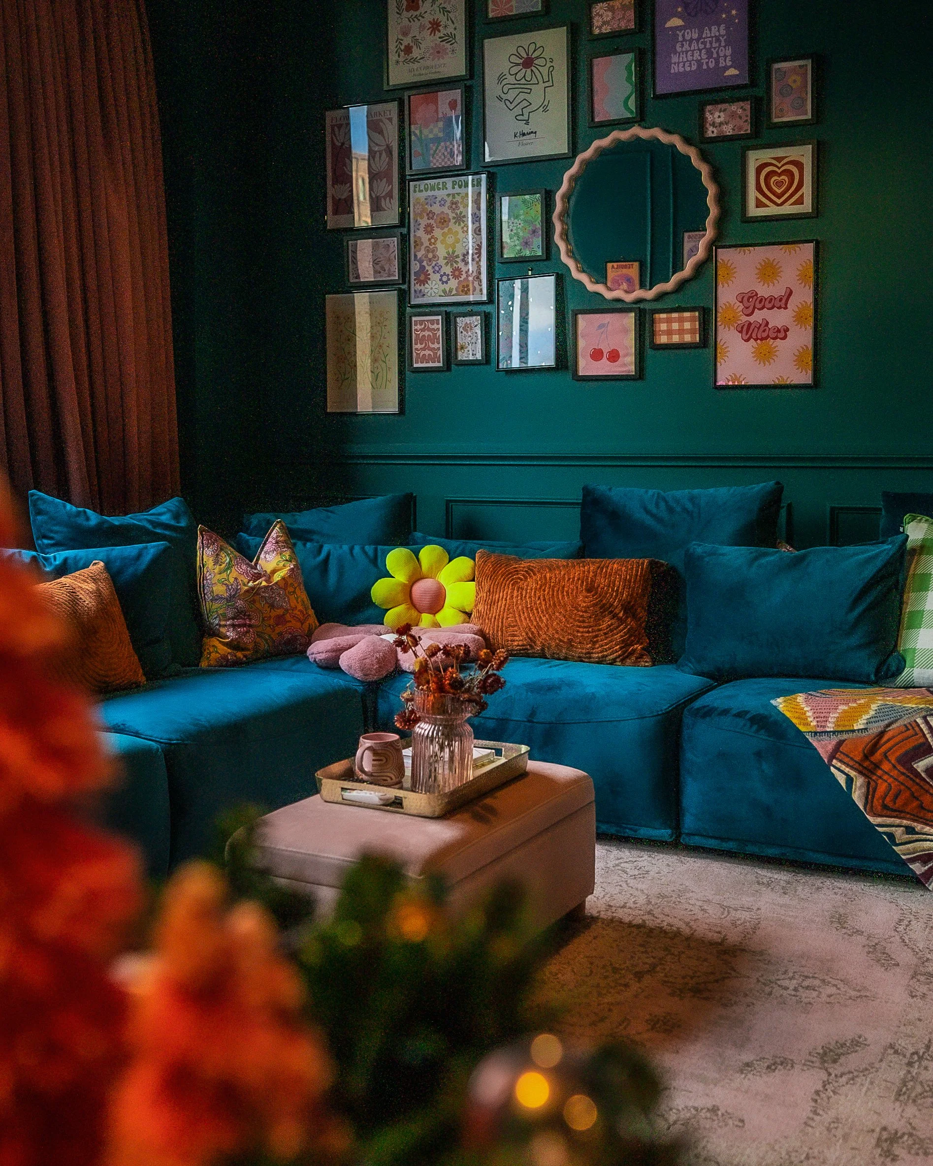 Cozy living room corner with a teal velvet sectional sofa decorated with colorful pillows, a pink ottoman with a tray holding a vase and mug, and a gallery wall with various framed art and a round mirror.