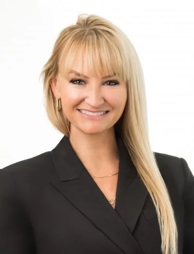 A woman with blonde hair, wearing a black blazer and gold jewelry, smiling against a white background.