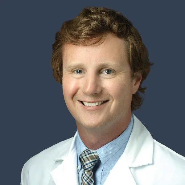 Portrait of a smiling male doctor with light brown hair, wearing a white coat, light blue shirt, and plaid tie, against a dark gray background.