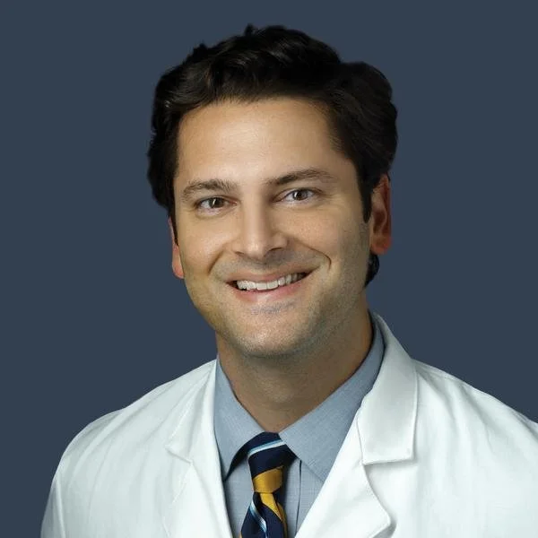 A smiling male doctor with dark hair, wearing a white coat, a light blue dress shirt, and a striped tie, against a dark gray background.
