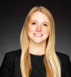 A young woman with long blonde hair smiling, wearing a black blazer over a black top, against a dark background.