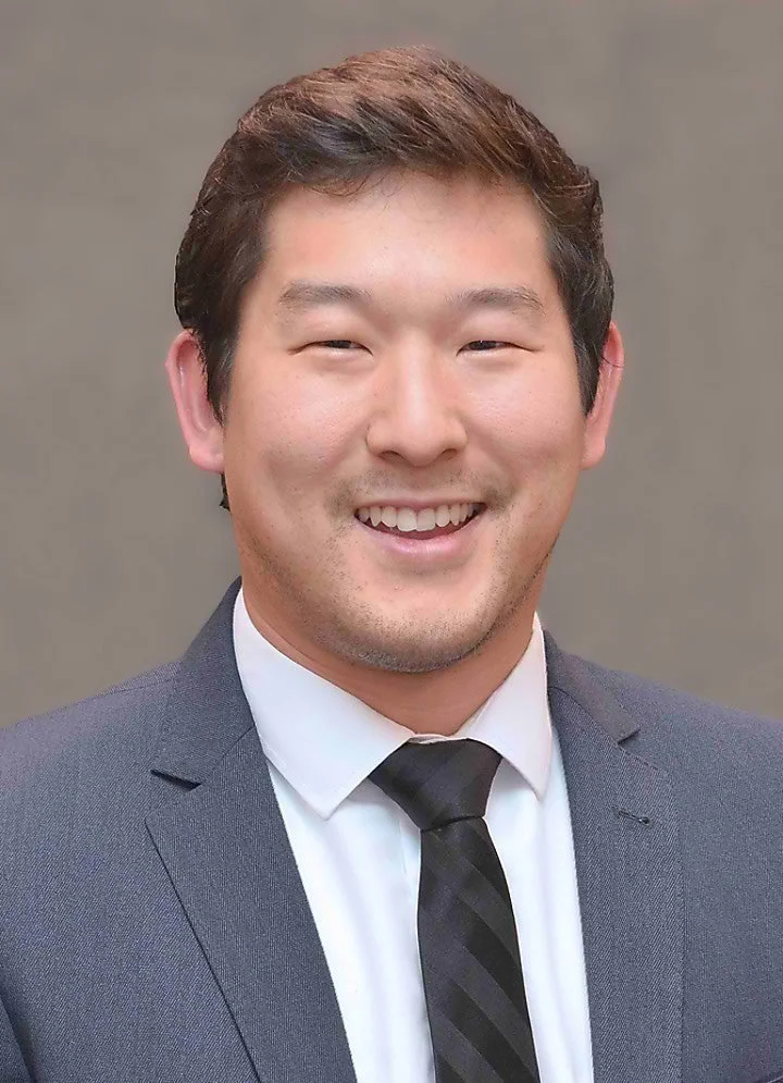 A smiling Asian man in a gray suit with a white shirt and black tie, posing for a portrait against a neutral background.