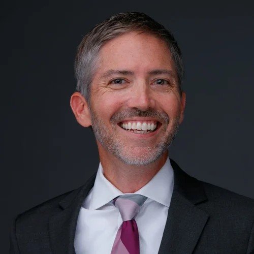 A smiling middle-aged man with gray hair and a beard wearing a black suit, white shirt, and purple tie, posing against a dark background.