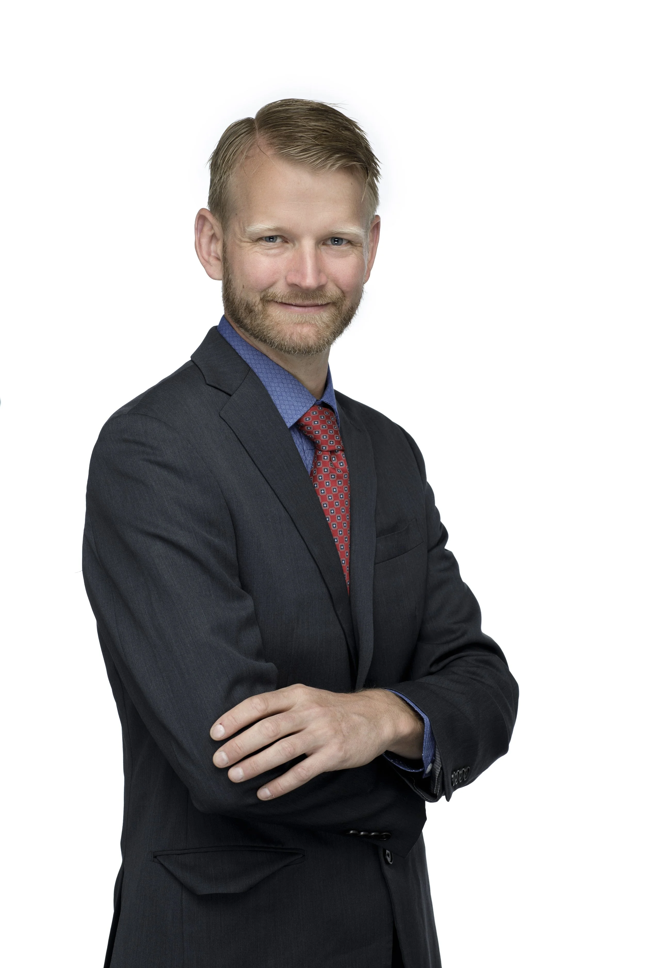 A man in a black suit with a red and blue tie, standing with his arms crossed, smiling, against a white background.