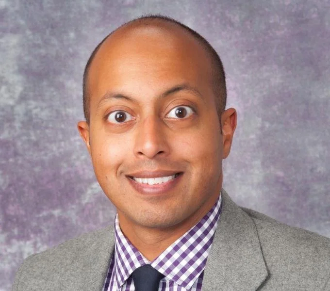 Headshot of a smiling man with a shaved head wearing a gray blazer, purple checkered shirt, and black tie, against a textured purple background.