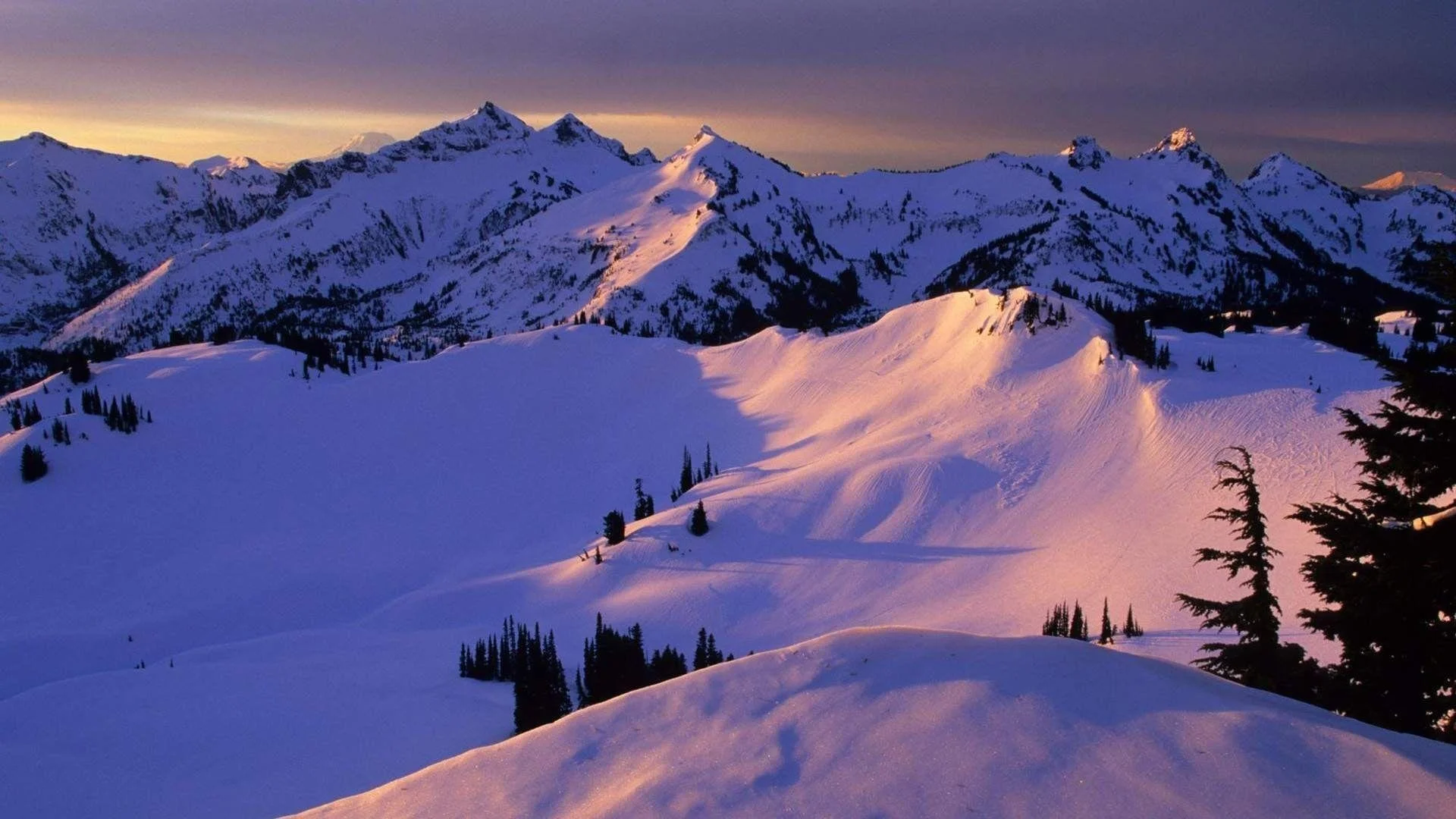 Snow-covered mountain range at sunset or sunrise with a colorful sky overhead, featuring peaks and ridges with some trees scattered across the landscape.