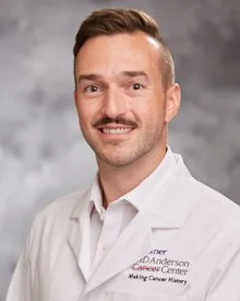 Portrait of a smiling man with a mustache, wearing a white medical coat with a name tag, in front of a gray studio background.