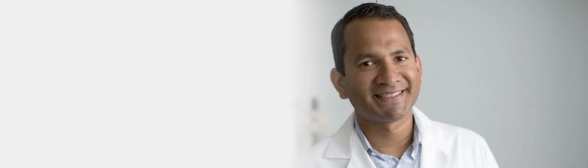 A smiling male doctor with short dark hair in a white lab coat standing against a light grey background.