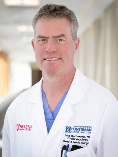 A male doctor in a white lab coat with university and hospital logos, smiling in a hospital or clinic hallway.