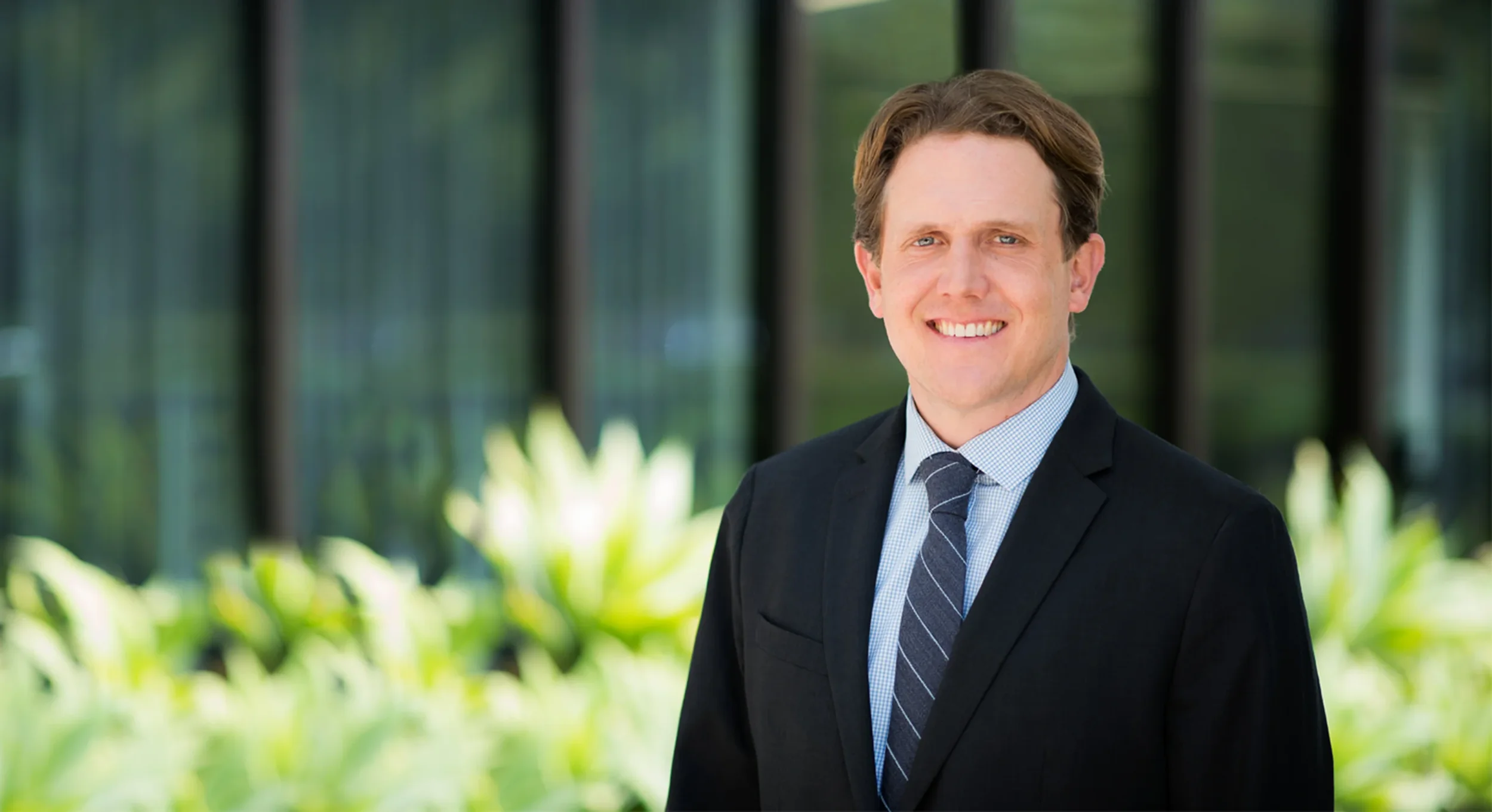 A smiling man in a black suit jacket, light blue shirt, and striped tie standing outdoors with a blurred background of glass windows and greenery.