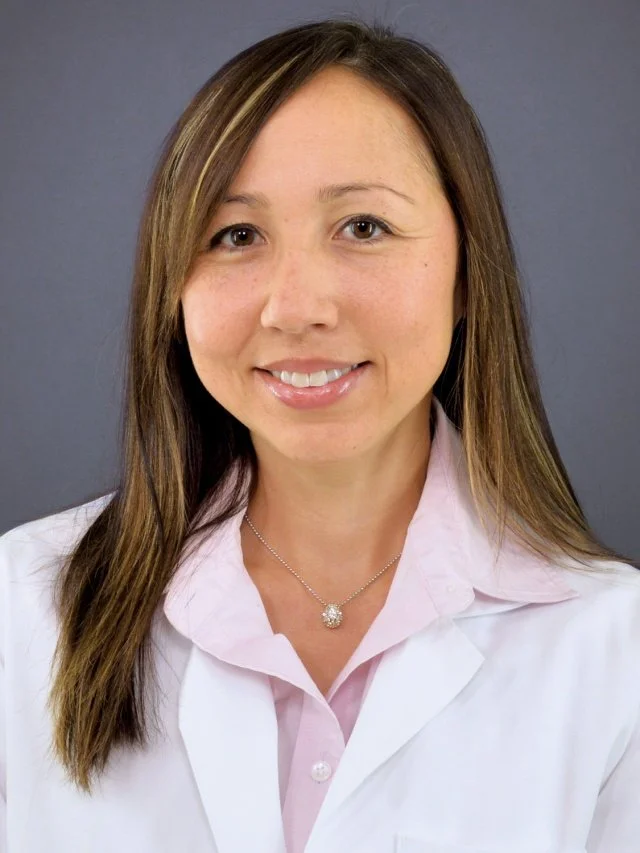 A woman with long brown hair, wearing a white lab coat and a pink shirt, smiling against a gray background.