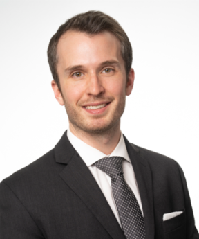 A man wearing a black suit, white shirt, and patterned tie, smiling against a plain white background.