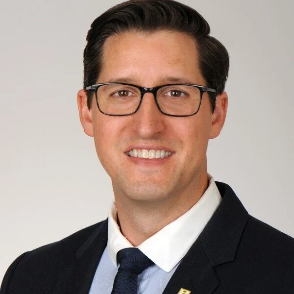 A man with glasses, wearing a dark suit, white shirt, and dark tie, smiling at the camera against a plain background.