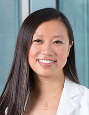 Portrait of a smiling woman with long dark hair, wearing a white lab coat, standing in front of a blue background.