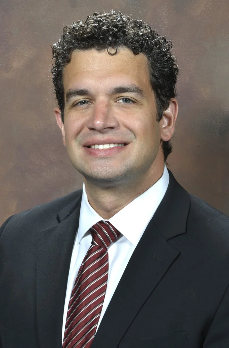 A professional headshot of a man with curly dark hair, wearing a black suit, white shirt, and a red striped tie, smiling at the camera against a brown background.