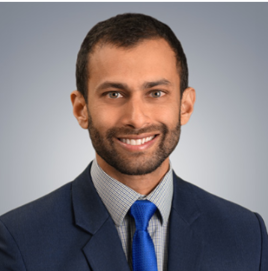Professional portrait of a smiling man with dark hair and beard, wearing a navy suit, light blue shirt, and blue tie, against a gray background.