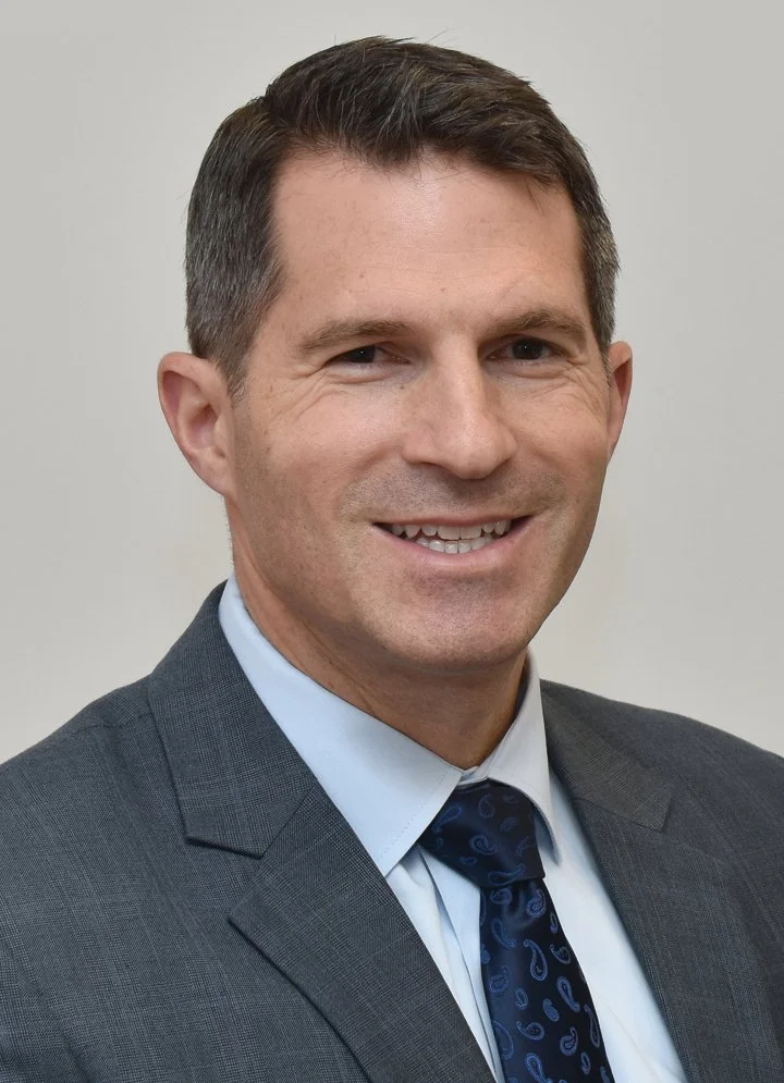 Headshot of a smiling man wearing a gray suit, light blue shirt, and dark patterned tie against a plain background.
