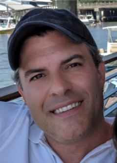 Close-up of a smiling man wearing a black cap, with boats and water in the background.