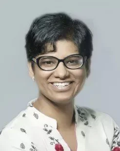 A woman with short dark hair wearing glasses and a white blouse, smiling against a plain background.