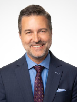 Portrait of a smiling man with dark hair and a beard, wearing a navy blue suit, light blue shirt, and patterned tie, against a white background.