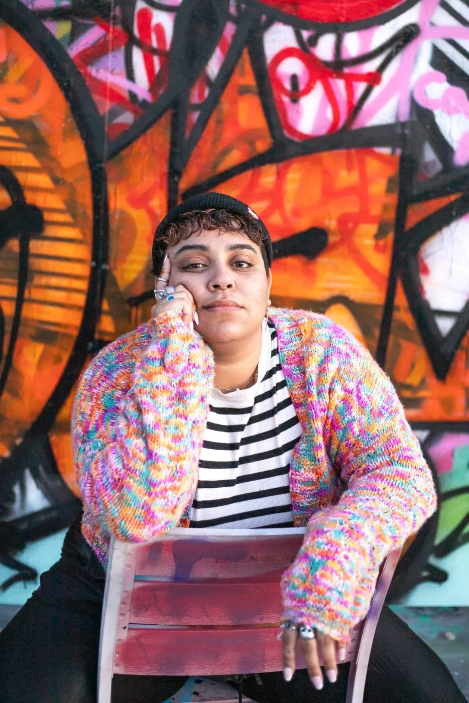Person with short curly hair wearing a black beanie, colorful sweater, and black and white striped shirt, sitting in front of a graffiti mural with vibrant orange, pink, and black colors, resting their head on their hand.