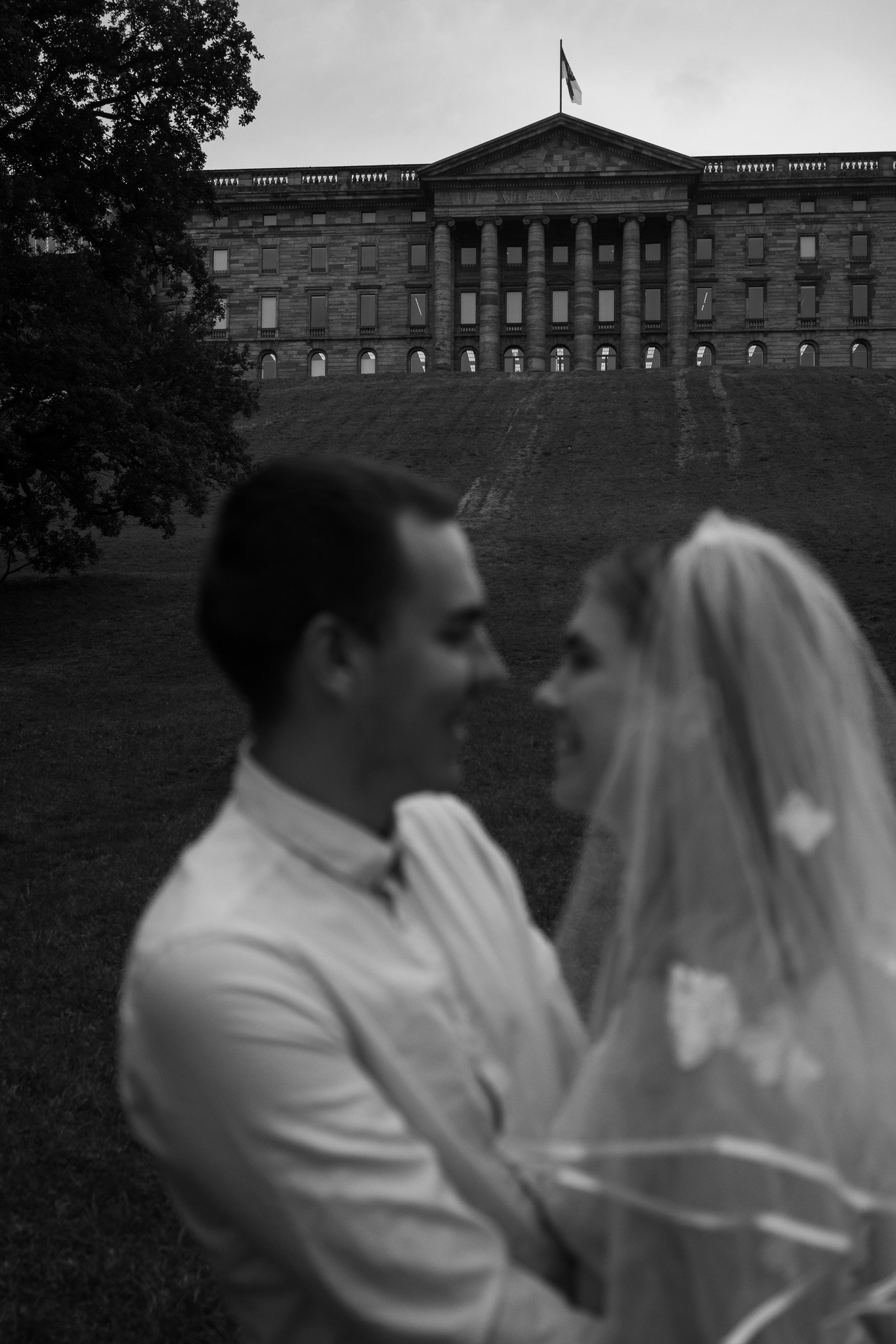 Ein Mann und eine Frau, vermutlich Braut und Bräutigam, stehen sich gegenüber im Freien mit der Reichstagsgebäudedemo im Hintergrund, in Schwarz-Weiß-Fotografie.