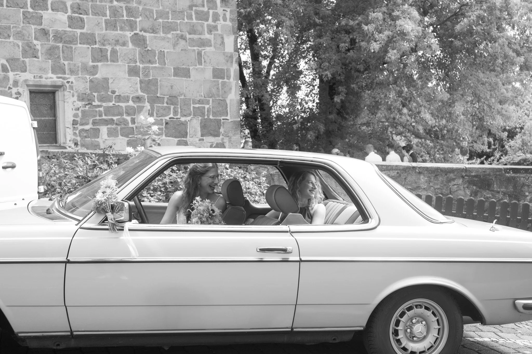 Zwei Frauen sitzen in einem klassischen Auto, es ist ein besonderer Anlass, vermutlich eine Hochzeit, mit Blumen am Auto. Sie lachen und scheinen fröhlich zu sein, während sie draußen unterwegs sind.