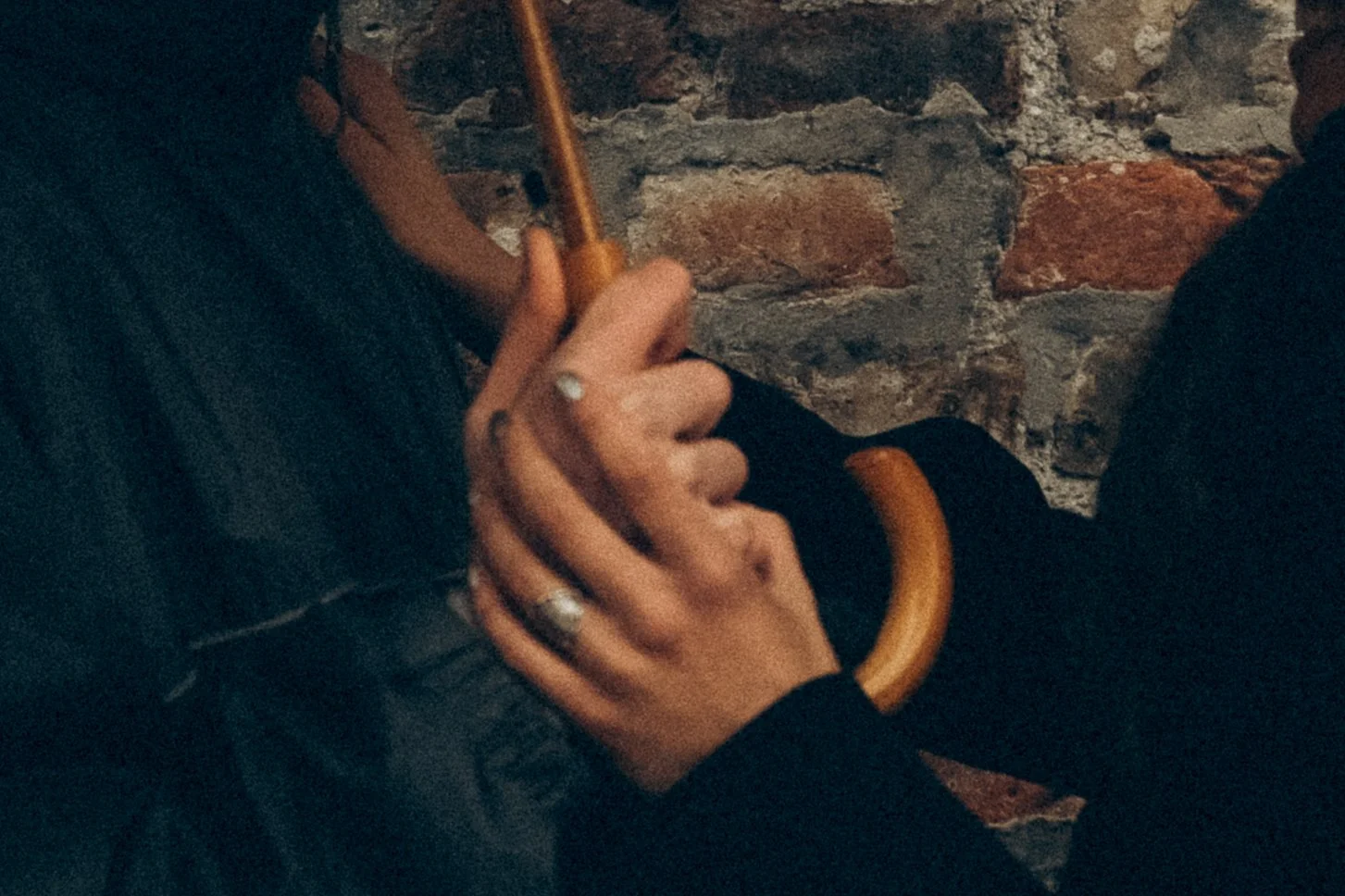 Person hält einen braunen Regenschirm und ein Smartphone in den Händen, vor einer Ziegelmauer.